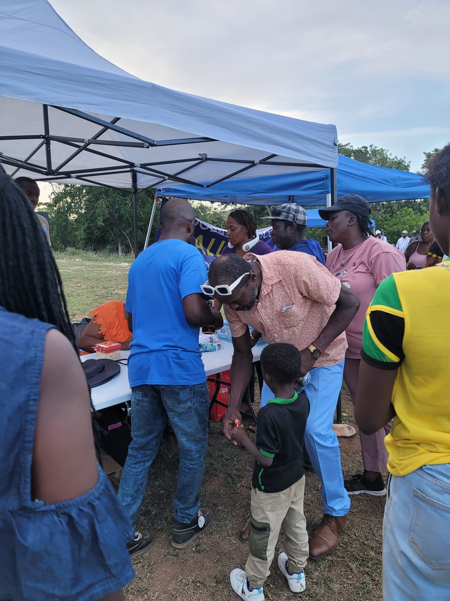 Community members gathered under a blue pop-up tent as volunteers sign students up and distribute school supplies at an outdoor Back 2 School event