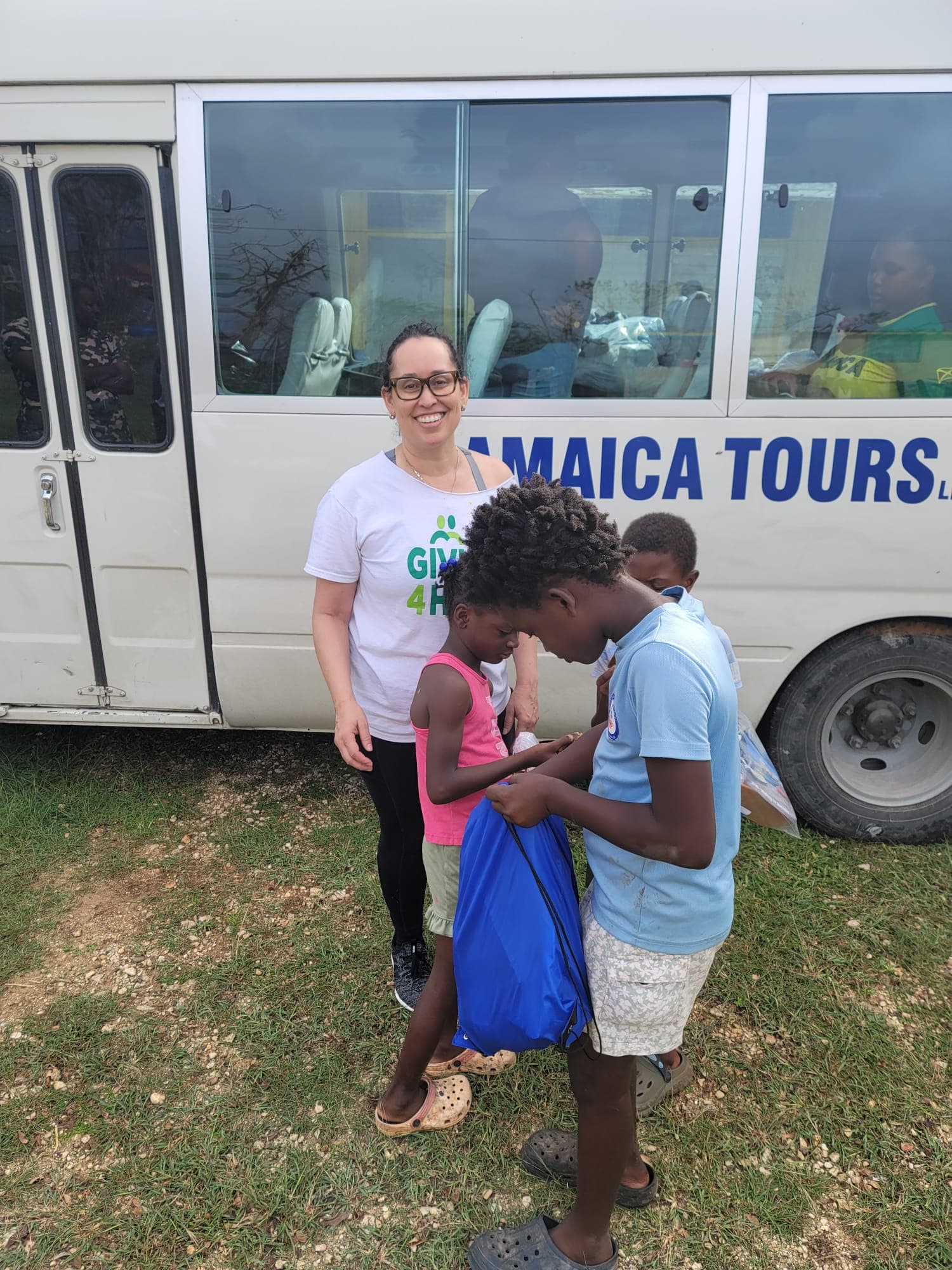 Volunteer in a Giving 4 Hope shirt handing a blue supply bag to children beside a Jamaica Tours Limited bus