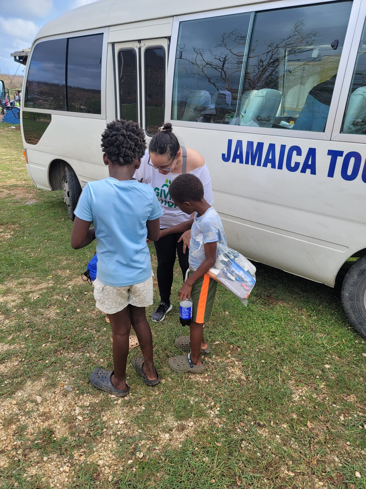 Volunteer at the Jamaica Tours bus handing drinking water and supplies to two children after the storm, damaged trees visible in the background