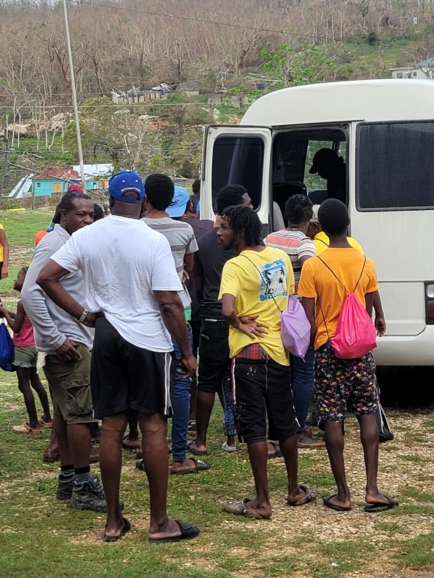 Community members waiting in line at the Jamaica Tours bus for relief supplies, with hurricane-damaged trees and homes visible behind