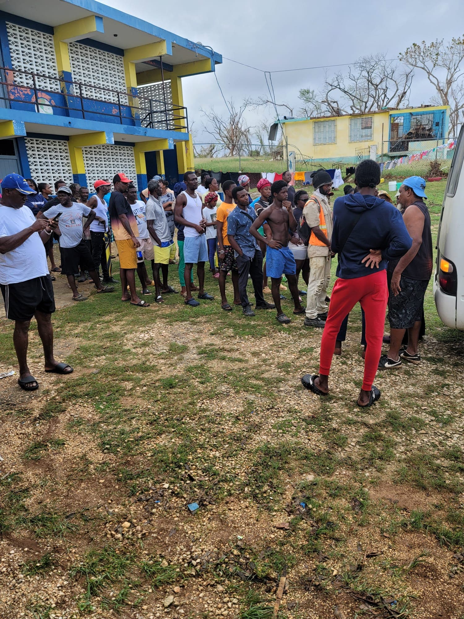 Large community gathering outside a school building awaiting distribution, overcast post-storm sky
