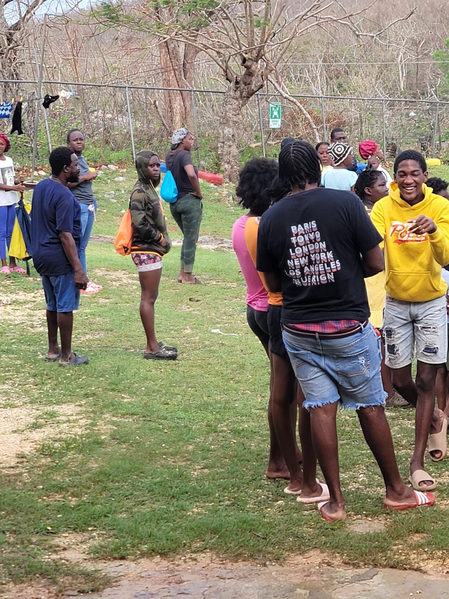 Community members waiting at an assembly-point distribution area with relief drawstring bags; stripped trees in the background show the storm's aftermath