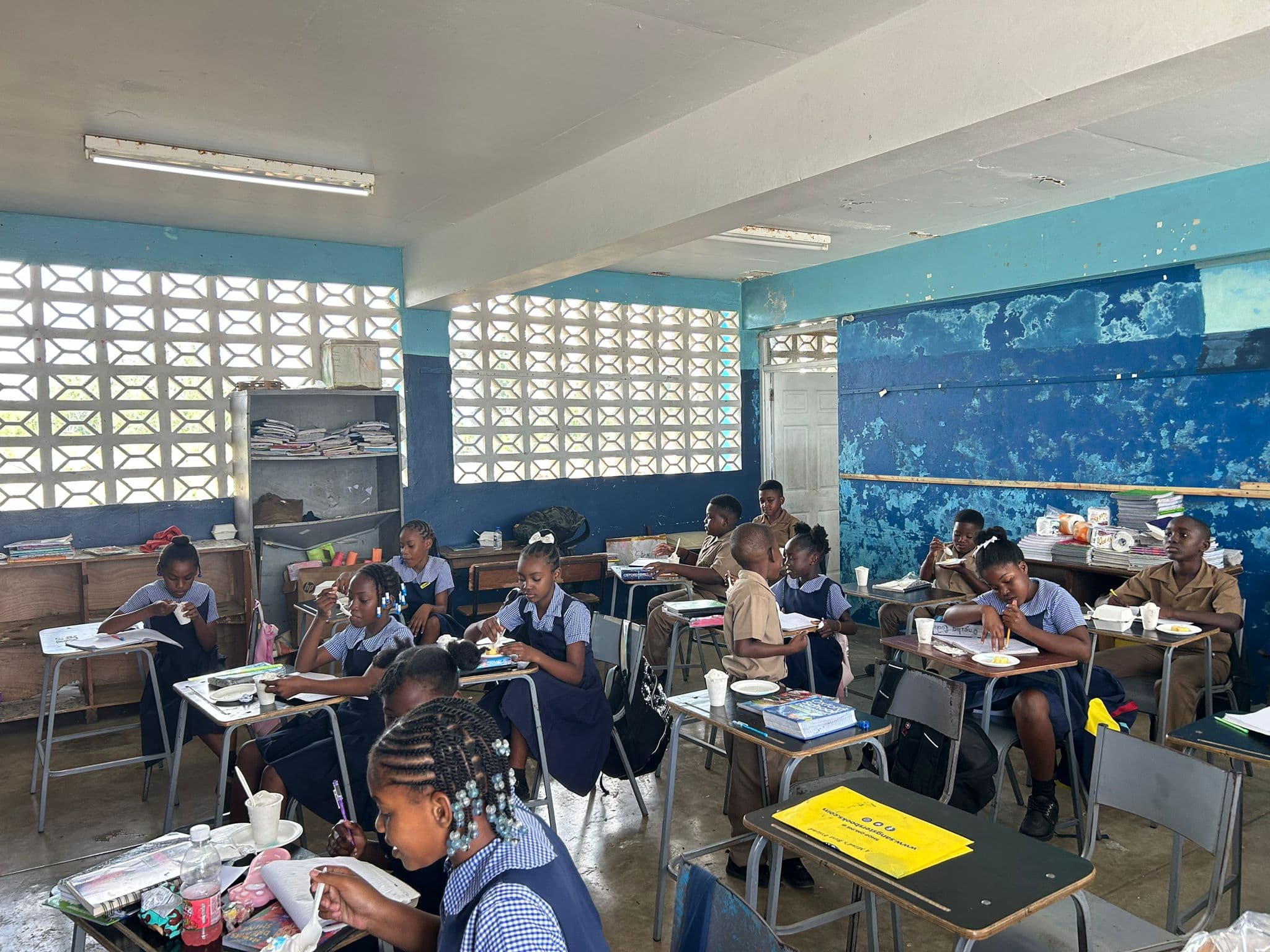 Wide view of a Jamaican primary classroom with blue walls and white breeze-block windows; students in tan uniforms sit at desks with cups of ice cream.