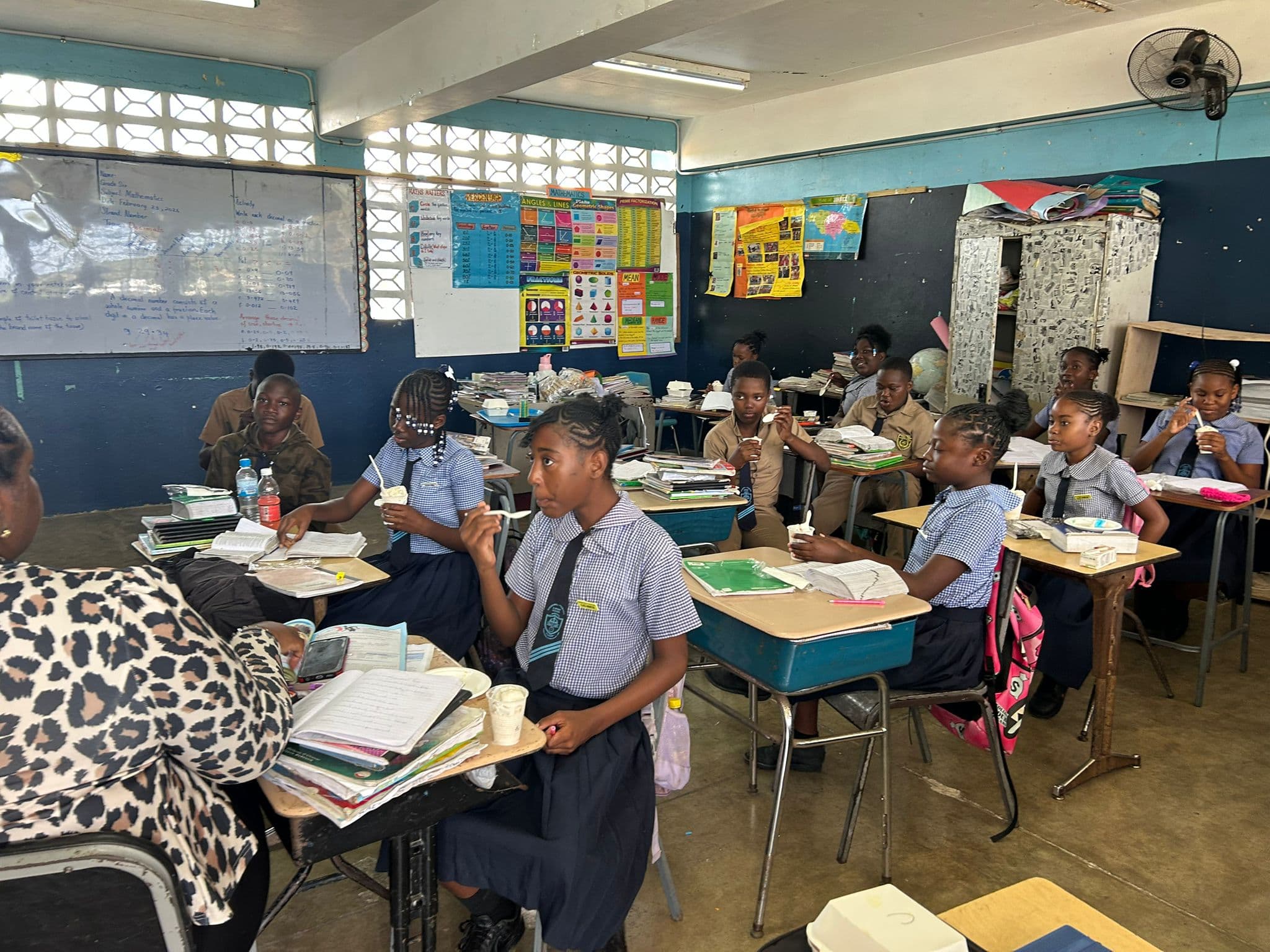 Students in white tops and navy pinafores eat ice cream at shared desk pods; colorful educational posters line the walls.