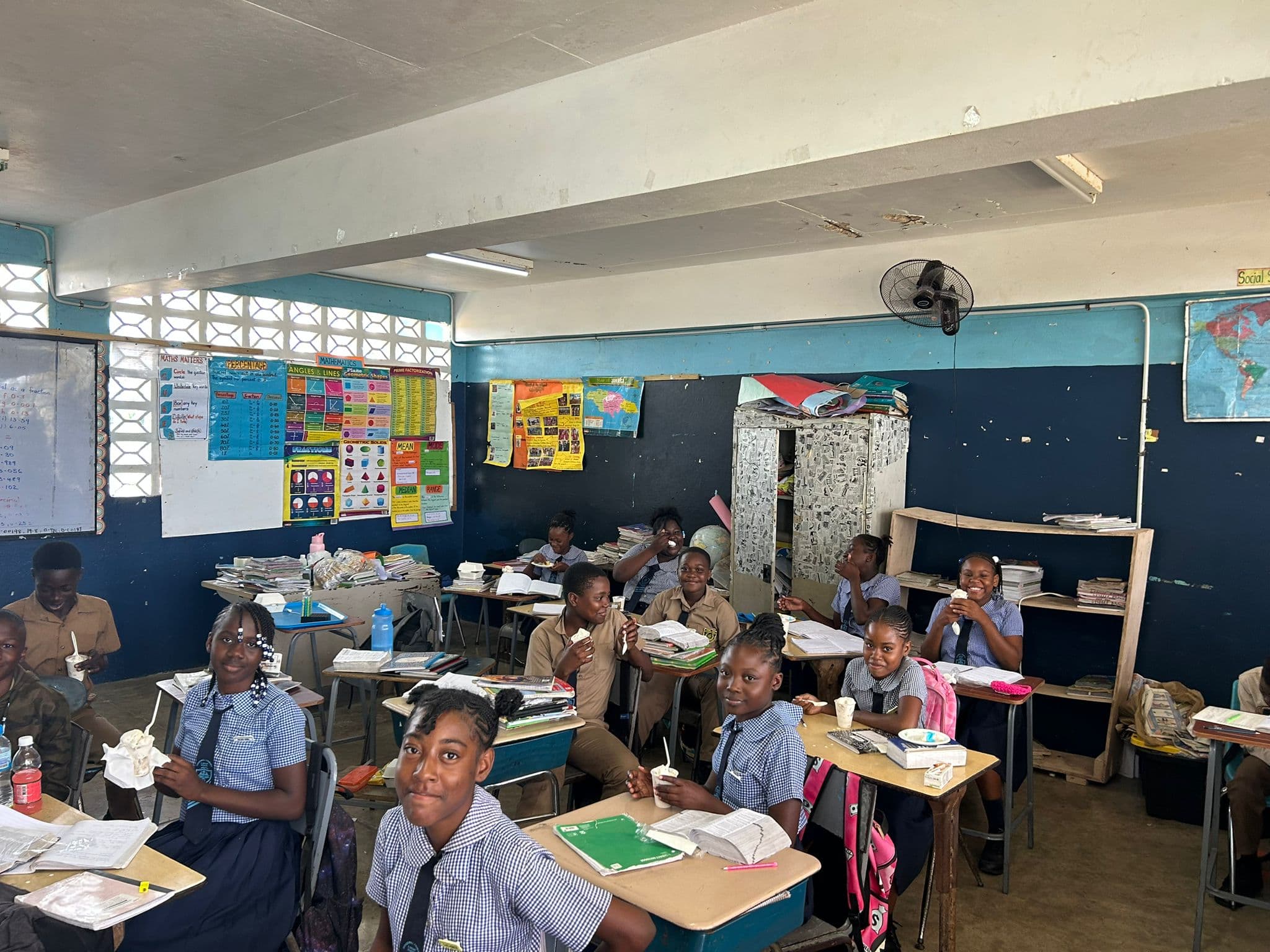 Students in navy pinafores and white tops smile and hold up ice cream cups in a classroom lined with educational posters.