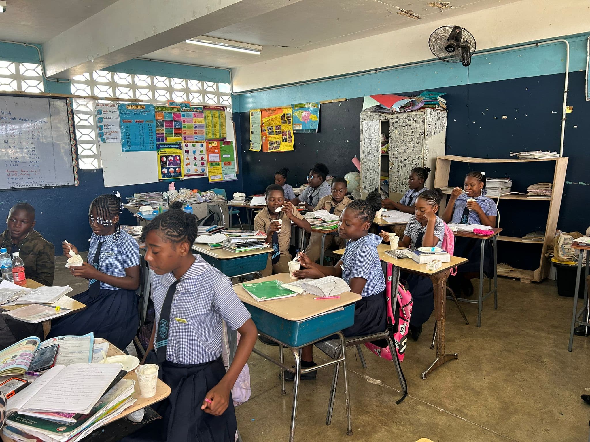 Young students look up from their schoolwork with ice cream cups in hand, a colorful classroom display behind them.